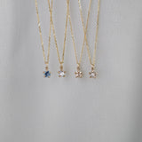 Four delicate yellow gold pendant necklaces by Laurie Fleming Jewellery draped over white fabric. From left to right: the blue-green teal Sapphire Moondew Necklace, the Rose Cut Mother of Pearl Moondew Necklace, the Rose Cut Diamond Moondew Necklace, and the Brilliant Cut Diamond Moondew Necklace. - Laurie Fleming Jewellery