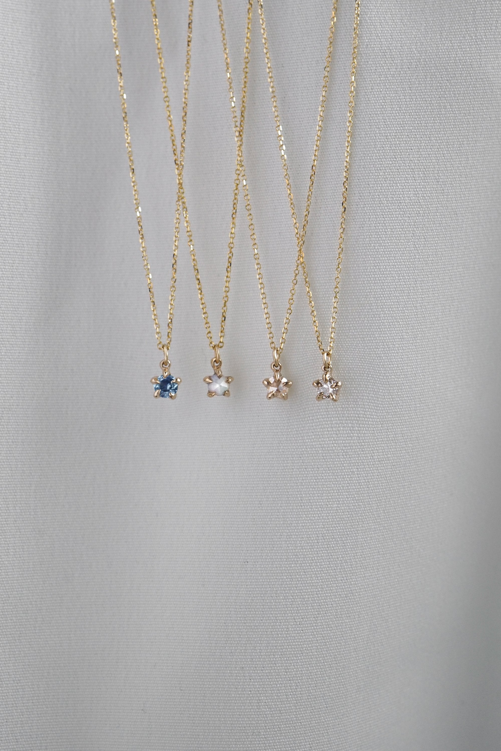 Four delicate yellow gold pendant necklaces by Laurie Fleming Jewellery draped over white fabric. From left to right: the blue-green teal Sapphire Moondew Necklace, the Rose Cut Mother of Pearl Moondew Necklace, the Rose Cut Diamond Moondew Necklace, and the Brilliant Cut Diamond Moondew Necklace. - Laurie Fleming Jewellery