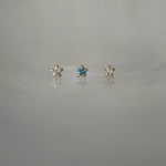 A photo of three Moondew stud earrings laid out in a row against a medium grey background. From left to right: the Round Brilliant Cut Diamond Moondew Stud, the Blue-Green Teal Sapphire Moondew Stud, and the Rose Cut Diamond Moondew Stud.