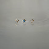 A photo of three Moondew stud earrings laid out in a row against a medium grey background. From left to right: the Round Brilliant Cut Diamond Moondew Stud, the Blue-Green Teal Sapphire Moondew Stud, and the Rose Cut Diamond Moondew Stud.