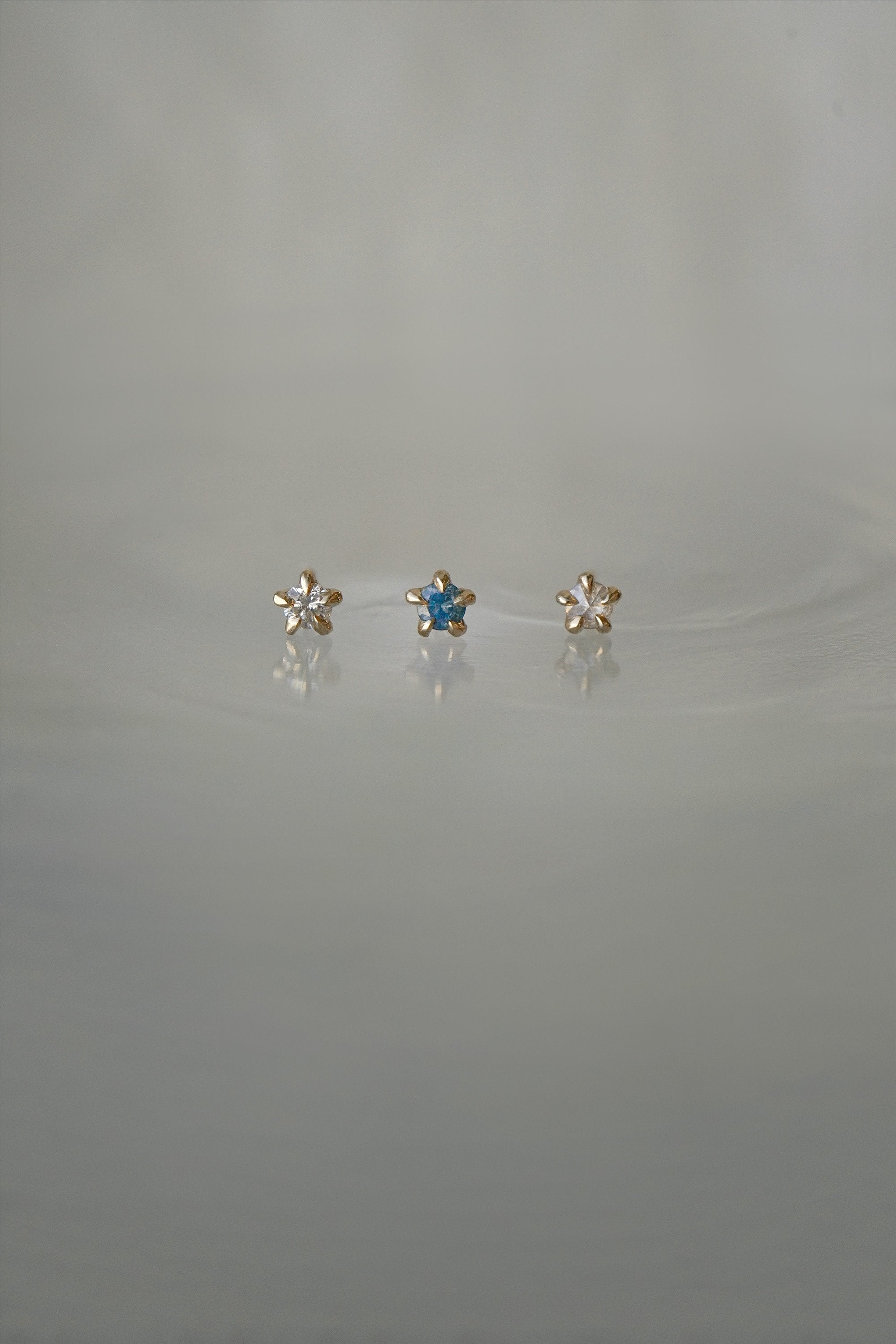 A photo of three Moondew stud earrings laid out in a row against a medium grey background. From left to right: the Round Brilliant Cut Diamond Moondew Stud, the Blue-Green Teal Sapphire Moondew Stud, and the Rose Cut Diamond Moondew Stud.
