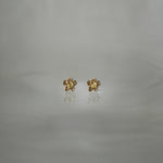 Two Laurie Fleming Jewellery "Buttercup Studs" resting on a medium grey background. The studs feature a hand-carved buttercup flower with a petite yellow sapphire centre.