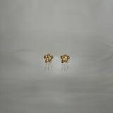 Two Laurie Fleming Jewellery "Buttercup Studs" resting on a medium grey background. The studs feature a hand-carved buttercup flower with a petite yellow sapphire centre.