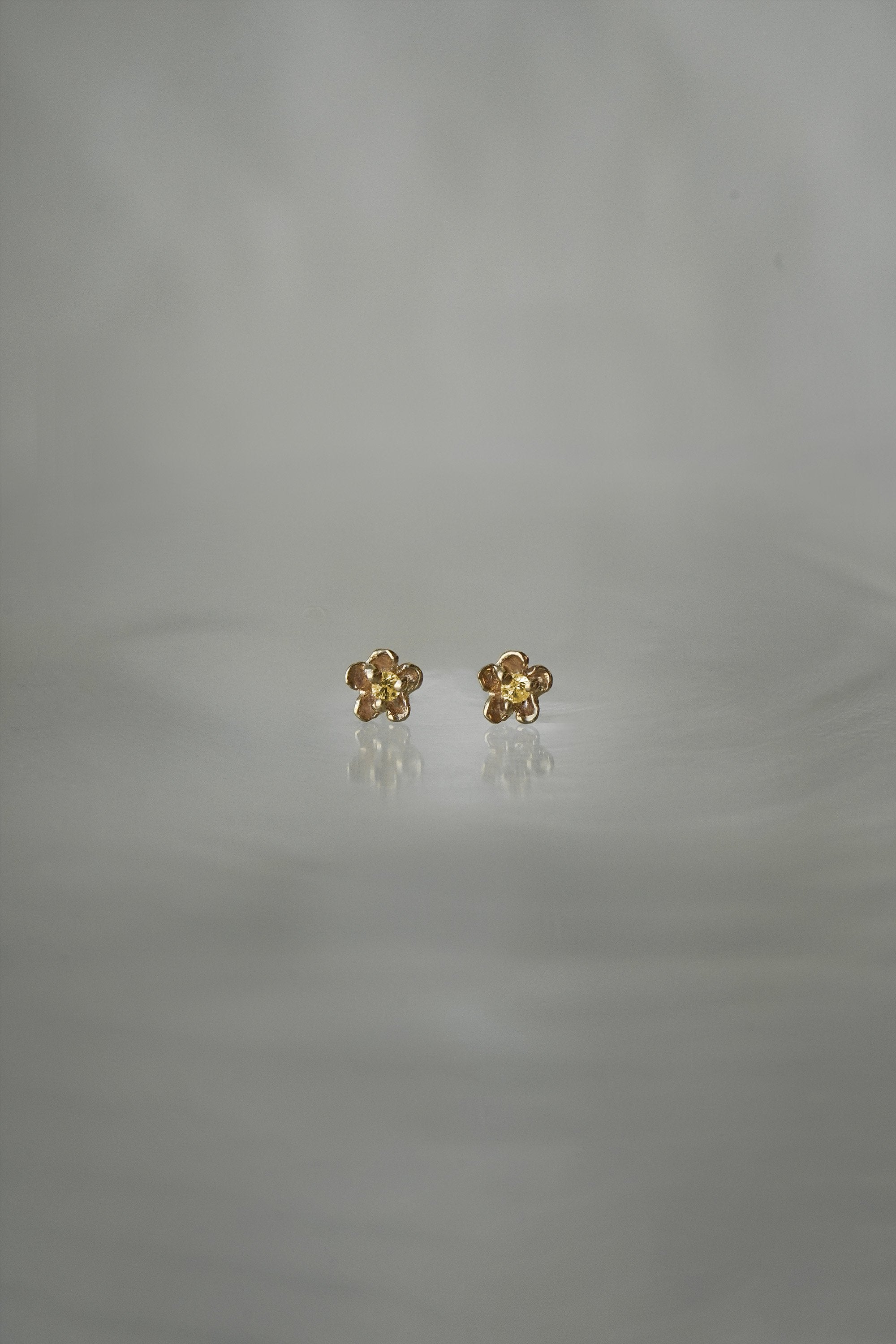 Two Laurie Fleming Jewellery "Buttercup Studs" resting on a medium grey background. The studs feature a hand-carved buttercup flower with a petite yellow sapphire centre.