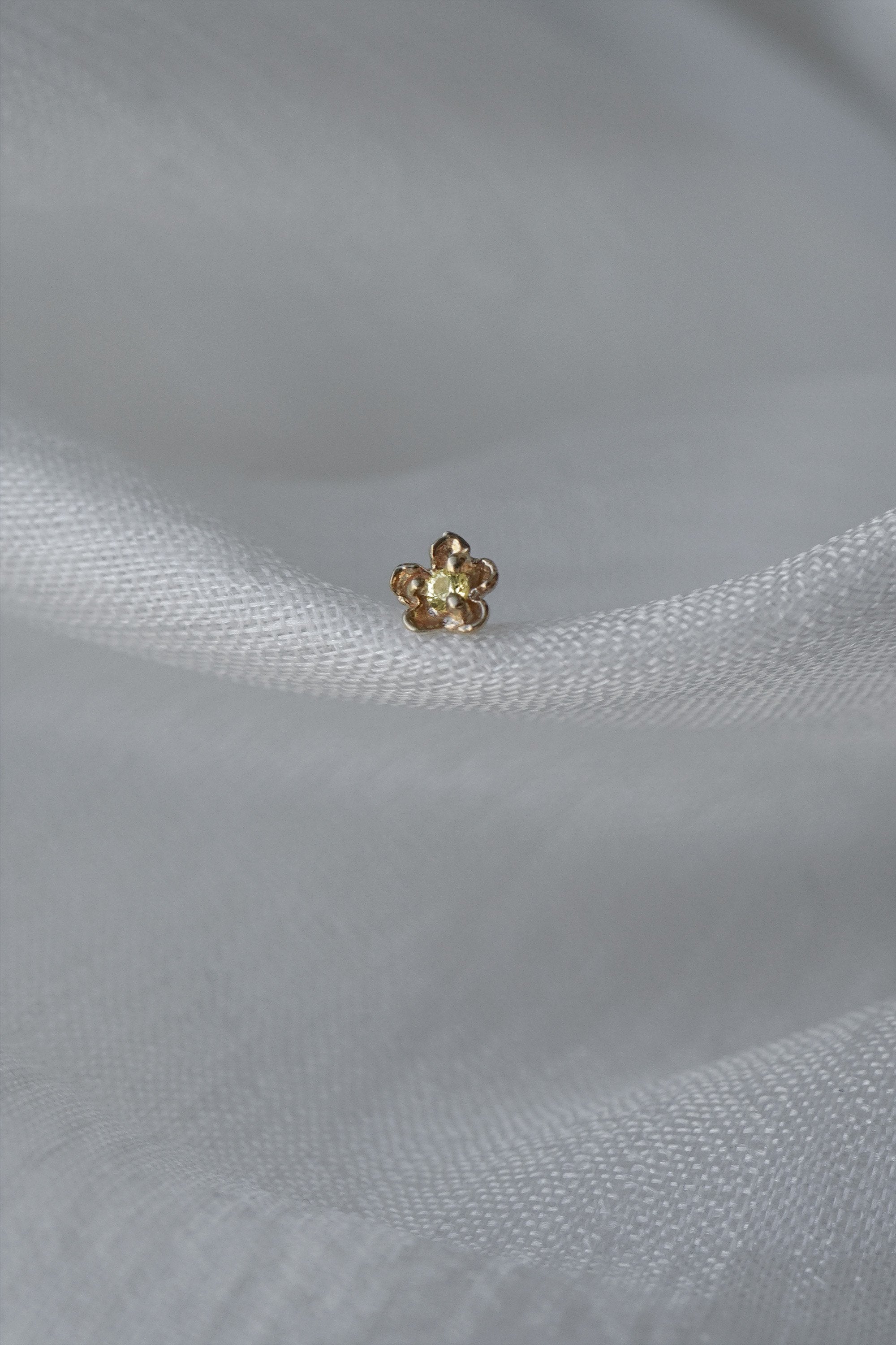 A single Buttercup Stud by Laurie Fleming Jewellery resting on loosely woven white silk. The earring features a hand-carved buttercup flower with a yellow sapphire centre.