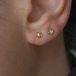 A close-up shot of an ear with two piercings wearing two petite studs by Laurie Fleming Jewellery. In the first hole, a "Buttercup Stud" with a hand-carved buttercup flower with a yellow sapphire, and in the second hole, a "Mother of Pearl Moondew Stud", with a rose cut mother of pearl stone held by five delicate claw-shaped prongs.