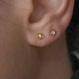 A close-up shot of an ear with two piercings wearing two petite studs by Laurie Fleming Jewellery. In the first hole, a "Buttercup Stud" with a hand-carved buttercup flower with a yellow sapphire, and in the second hole, a "Mother of Pearl Moondew Stud", with a rose cut mother of pearl stone held by five delicate claw-shaped prongs.