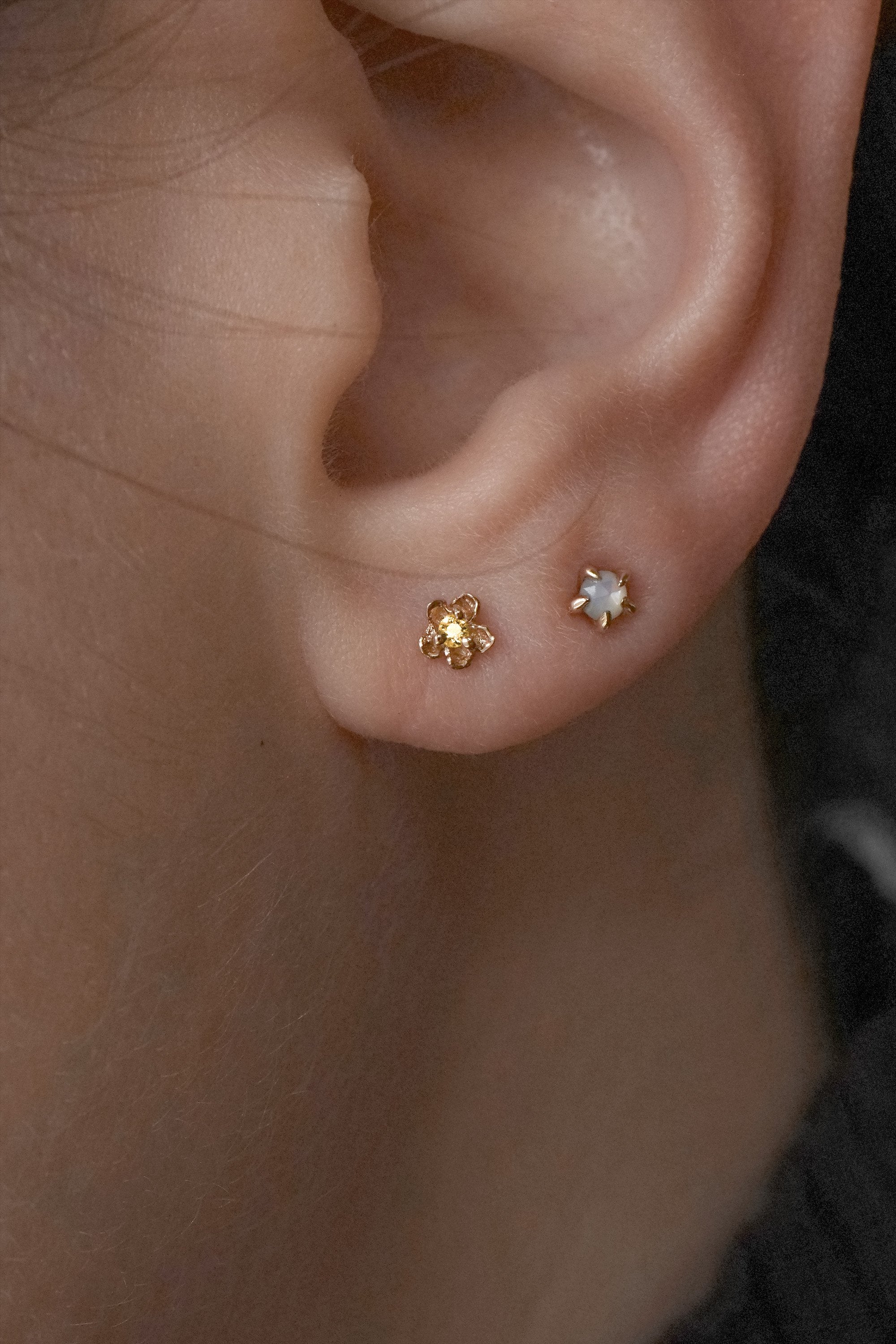 A close-up shot of an ear with two piercings wearing two petite studs by Laurie Fleming Jewellery. In the first hole, a "Buttercup Stud" with a hand-carved buttercup flower with a yellow sapphire, and in the second hole, a "Mother of Pearl Moondew Stud", with a rose cut mother of pearl stone held by five delicate claw-shaped prongs.