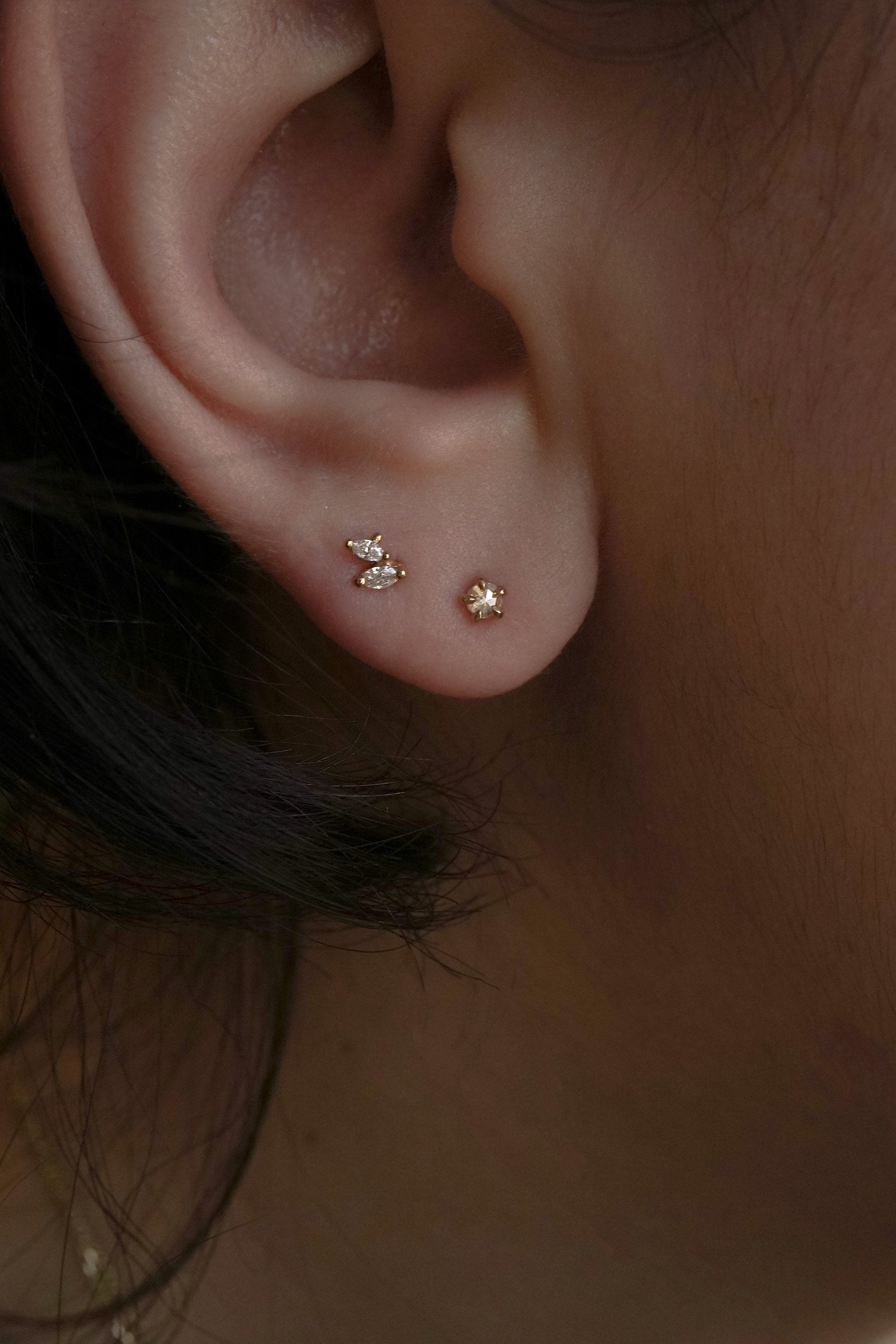 A close up of an ear wearing two solid gold Laurie Fleming Jewellery stud earrings. In the top piercing is a diamond Gossamer Wing stud featuring two small marquise cut diamonds. Below, the Rose Cut Diamond Moondew Stud.