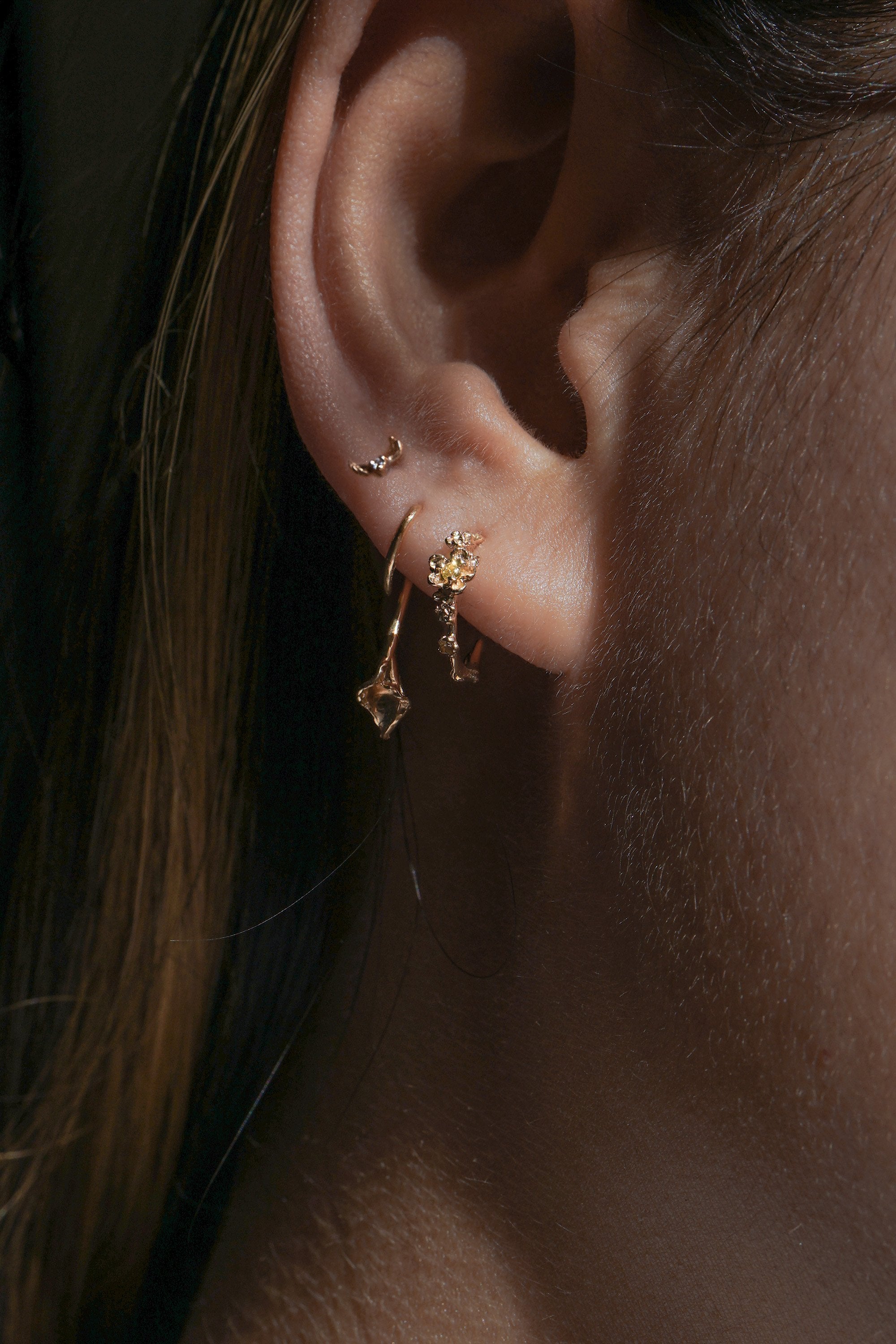 A close-up of an ear wearing three Laurie Fleming Jewellery earrings. In the first hole, a small buttercup hoop featuring handcarved buttercup flowers with a yellow sapphire centre. In the second hole, a Calla Lily earring, with a delicately hand-carved petite calla lily flower with a stem wrapping around the lobe. In the third hole, the tiniest hidden fairy stud with two delicate golden wings over a sparkling diamond.
