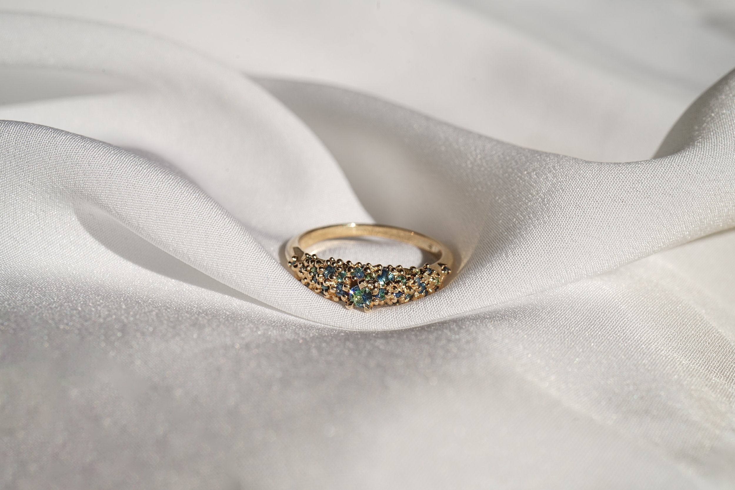 Laurie Fleming Jewellery's "Moss Signet Ring," a bold solid gold signet-style ring encrusted with varying shades and sizes of blue-green teal sapphires on the band. The ring is nestled in folds of smooth white fabric.