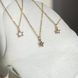 Three delicate yellow gold pendant necklaces by Laurie Fleming Jewellery draped upon the petal of a white flower. From left to right: the Rose Cut Mother of Pearl Moondew Necklace, the blue-green teal Sapphire Moondew Necklace, and the Rose Cut Diamond Moondew Necklace.