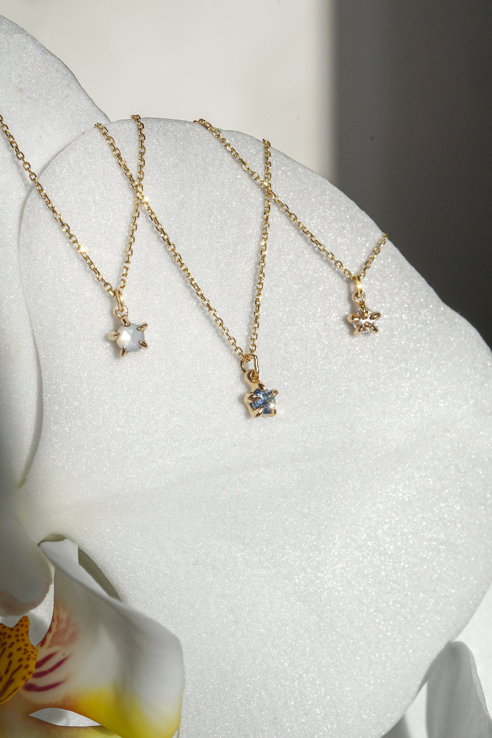 Three delicate yellow gold pendant necklaces by Laurie Fleming Jewellery draped upon the petal of a white flower. From left to right: the Rose Cut Mother of Pearl Moondew Necklace, the blue-green teal Sapphire Moondew Necklace, and the Rose Cut Diamond Moondew Necklace.