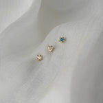 A photo of three moondew studs nestled in white, raw silk. From left to right, there is a rose cut diamond Moondew Stud, a round brilliant cut diamond Moondew stud, and a teal sapphire moondew stud. All three studs are in solid yellow gold.