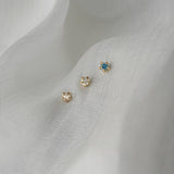 A photo of three moondew studs nestled in white, raw silk. From left to right, there is a rose cut diamond Moondew Stud, a round brilliant cut diamond Moondew stud, and a teal sapphire moondew stud. All three studs are in solid yellow gold.