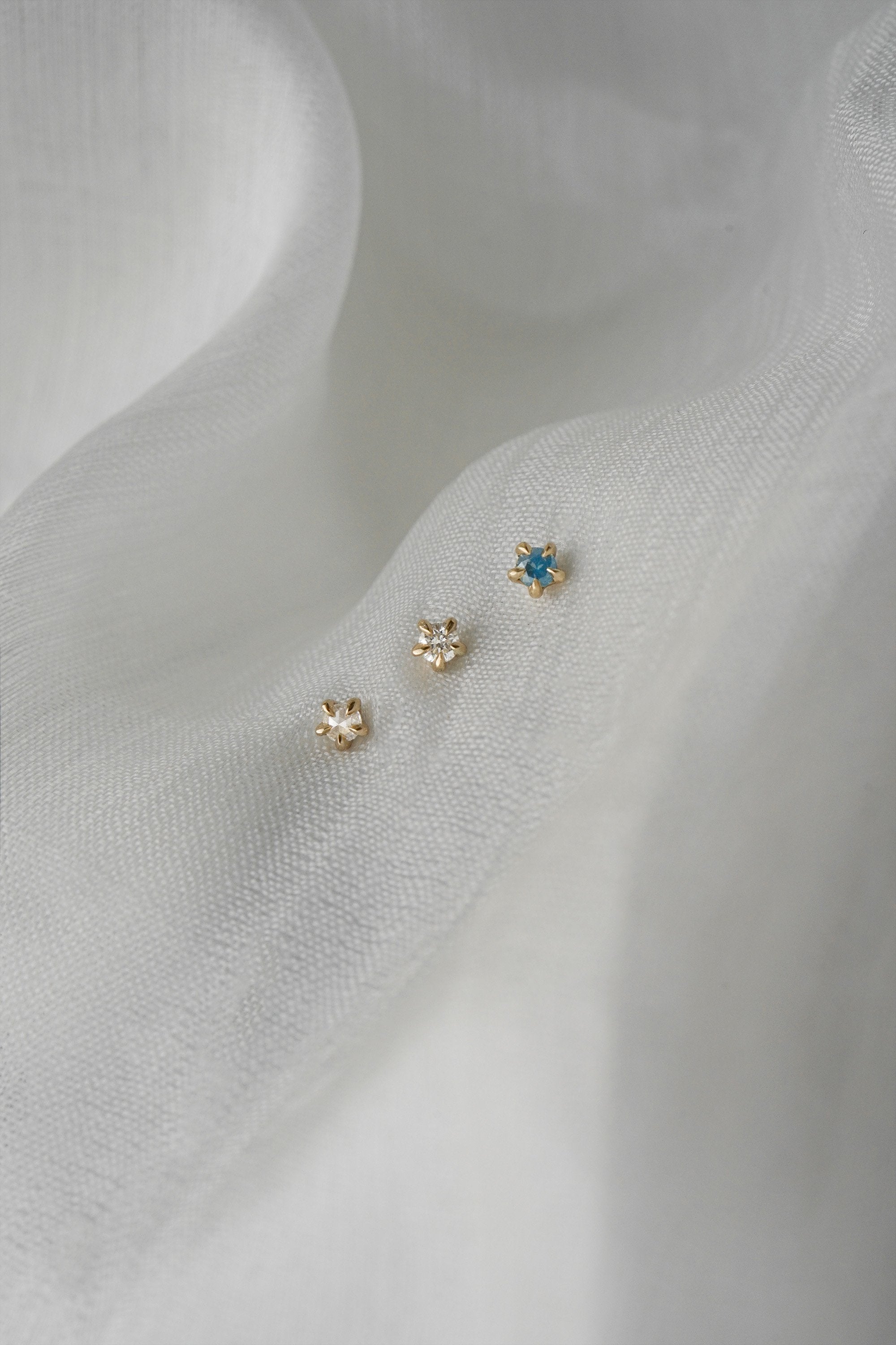 A photo of three moondew studs nestled in white, raw silk. From left to right, there is a rose cut diamond Moondew Stud, a round brilliant cut diamond Moondew stud, and a teal sapphire moondew stud. All three studs are in solid yellow gold.