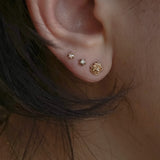 A photo of three Laurie Fleming Jewellery studs worn on an ear. From top to bottom, there is a rose cut diamond Moondew Stud, a round brilliant cut diamond Moondew stud, and a diamond dewdrop rose stud. All three studs are in solid yellow gold.