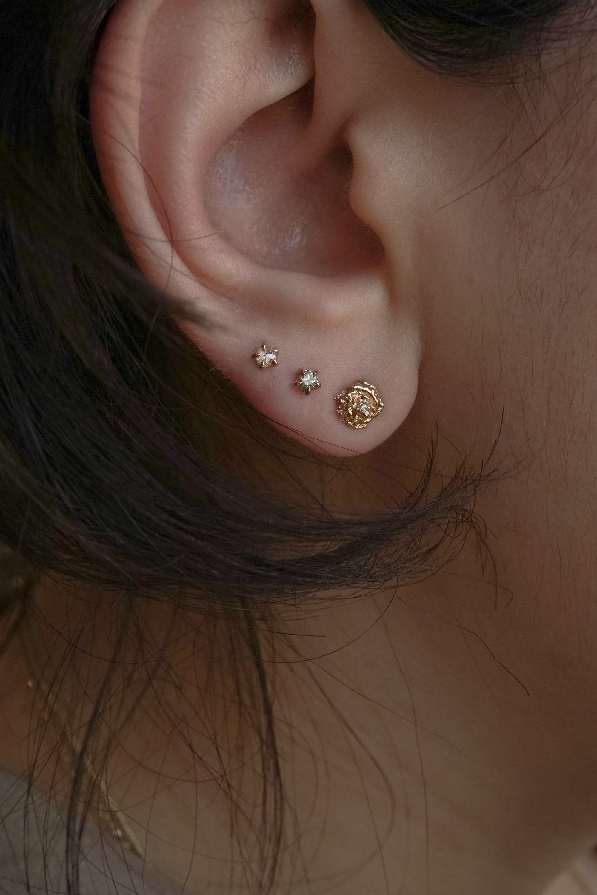 A photo of three Laurie Fleming Jewellery studs worn on an ear. From top to bottom, there is a rose cut diamond Moondew Stud, a round brilliant cut diamond Moondew stud, and a diamond dewdrop rose stud. All three studs are in solid yellow gold.