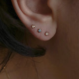A close up of an ear wearing three Laurie Fleming Jewellery studs. On top is the rose cut diamond Moondew stud. In the middle is a blue-green teal Sapphire Moondew Stud. At the bottom is a Brilliant Cut Diamond Moondew Stud.
