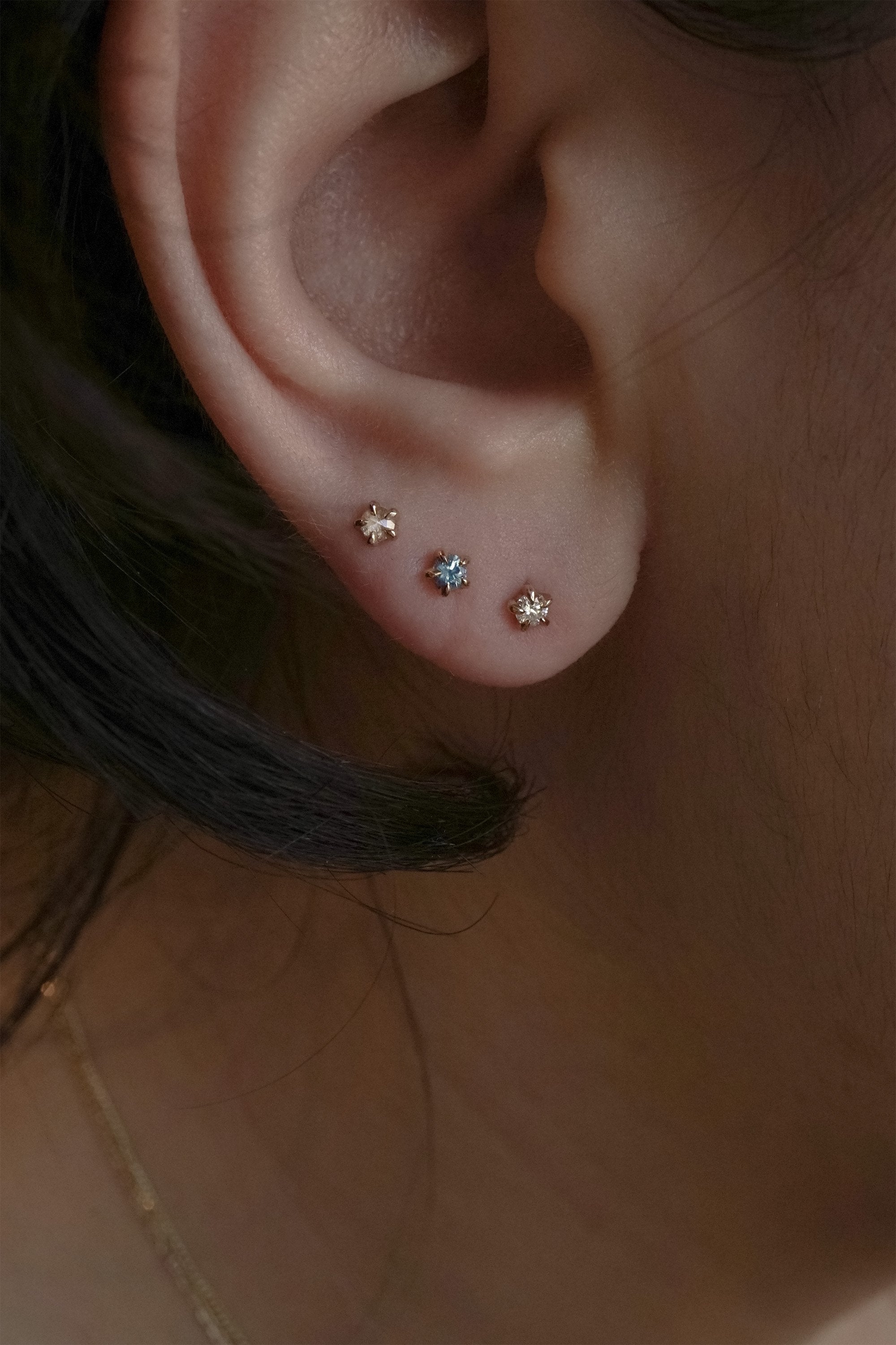A close up of an ear wearing three Laurie Fleming Jewellery studs. On top is the rose cut diamond Moondew stud. In the middle is a blue-green teal Sapphire Moondew Stud. At the bottom is a Brilliant Cut Diamond Moondew Stud.
