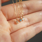 Three delicate yellow gold pendant necklaces by Laurie Fleming Jewellery held draped over the fingers of a hand. From left to right: the Rose Cut Diamond Moondew Necklace, the blue-green teal Sapphire Moondew Necklace, and the Rose Cut Mother of Pearl Moondew Necklace.