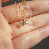Three delicate yellow gold pendant necklaces by Laurie Fleming Jewellery held draped over the fingers of a hand. From left to right: the Rose Cut Diamond Moondew Necklace, the blue-green teal Sapphire Moondew Necklace, and the Rose Cut Mother of Pearl Moondew Necklace.