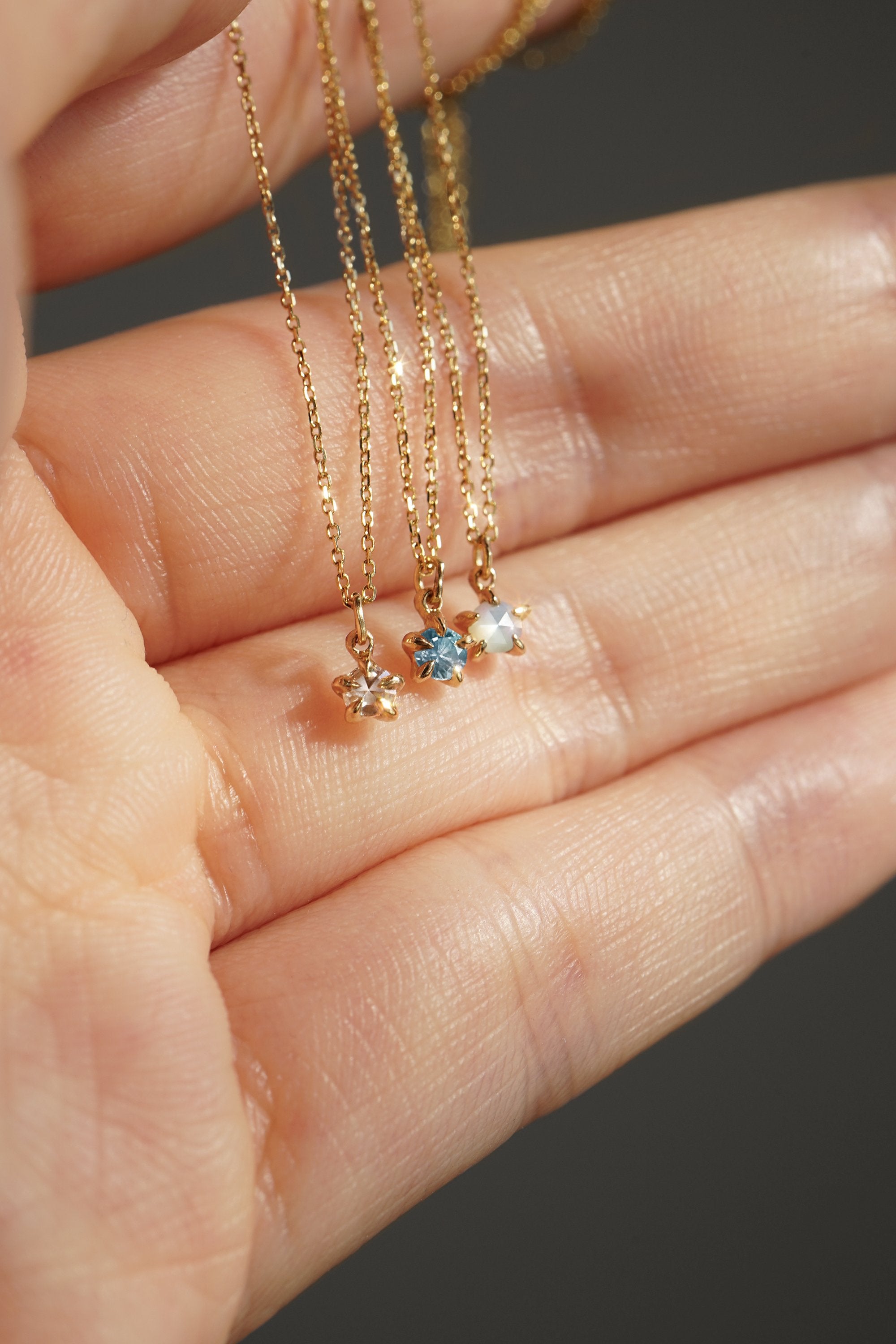 Three delicate yellow gold pendant necklaces by Laurie Fleming Jewellery held draped over the fingers of a hand. From left to right: the Rose Cut Diamond Moondew Necklace, the blue-green teal Sapphire Moondew Necklace, and the Rose Cut Mother of Pearl Moondew Necklace.