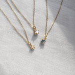 Three delicate yellow gold pendant necklaces by Laurie Fleming Jewellery draped over white raw silk. From left to right: the Rose Cut Diamond Moondew Necklace, the Rose Cut Mother of Pearl Moondew Necklace, and the blue-green teal Sapphire Moondew Necklace.
