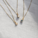 Three delicate yellow gold pendant necklaces by Laurie Fleming Jewellery draped over white raw silk. From left to right: the Rose Cut Diamond Moondew Necklace, the Rose Cut Mother of Pearl Moondew Necklace, and the blue-green teal Sapphire Moondew Necklace.