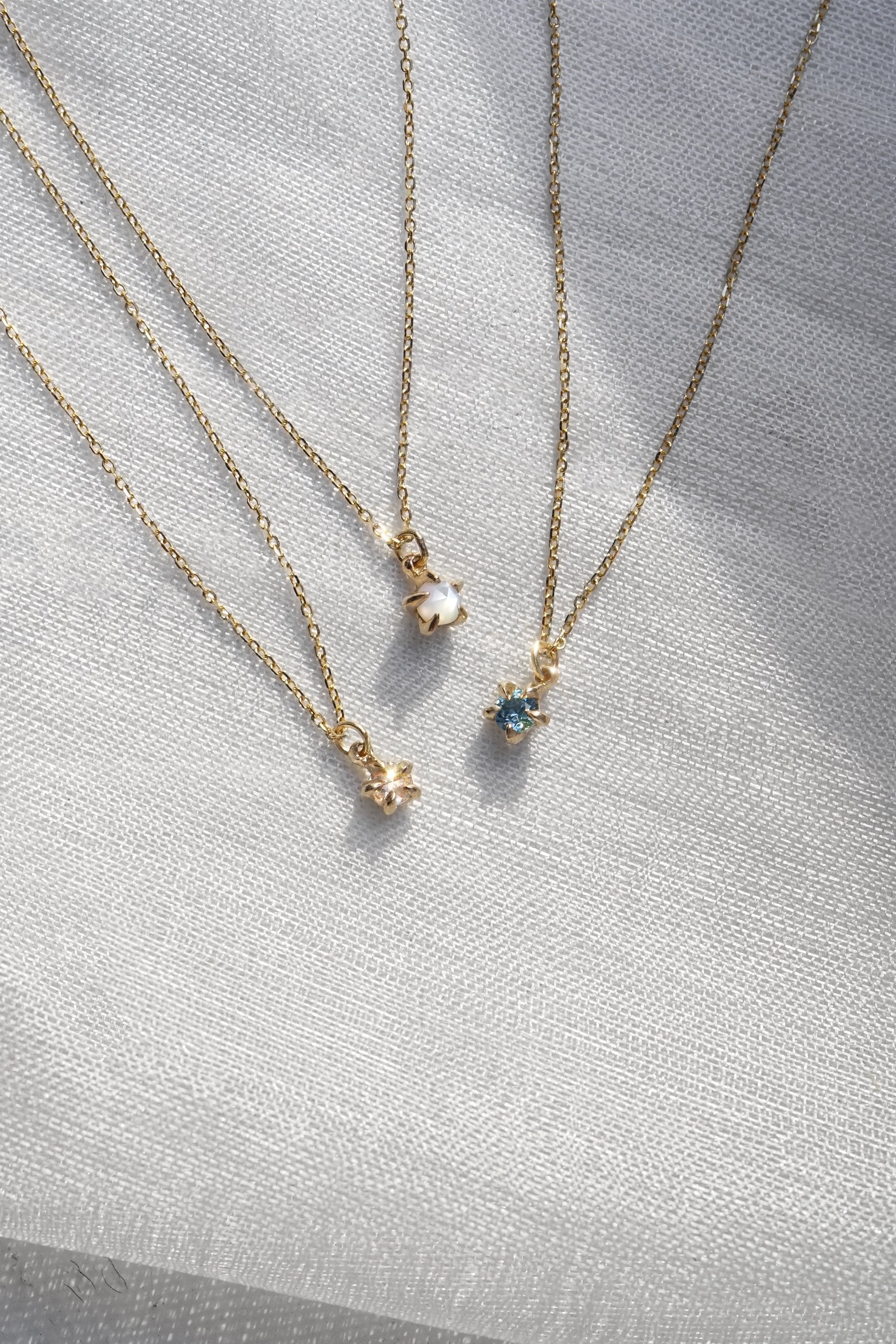Three delicate yellow gold pendant necklaces by Laurie Fleming Jewellery draped over white raw silk. From left to right: the Rose Cut Diamond Moondew Necklace, the Rose Cut Mother of Pearl Moondew Necklace, and the blue-green teal Sapphire Moondew Necklace.