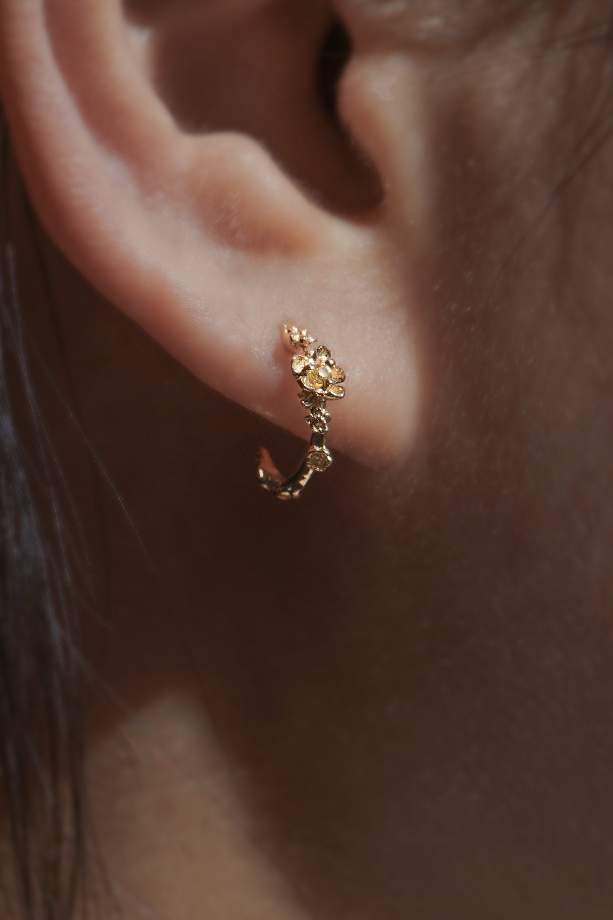 Close-up of an ear wearing a Laurie Fleming Jewellery "Small Buttercup Hoop" earring, featuring petite hand-carved buttercup flowers with a yellow sapphire centre.