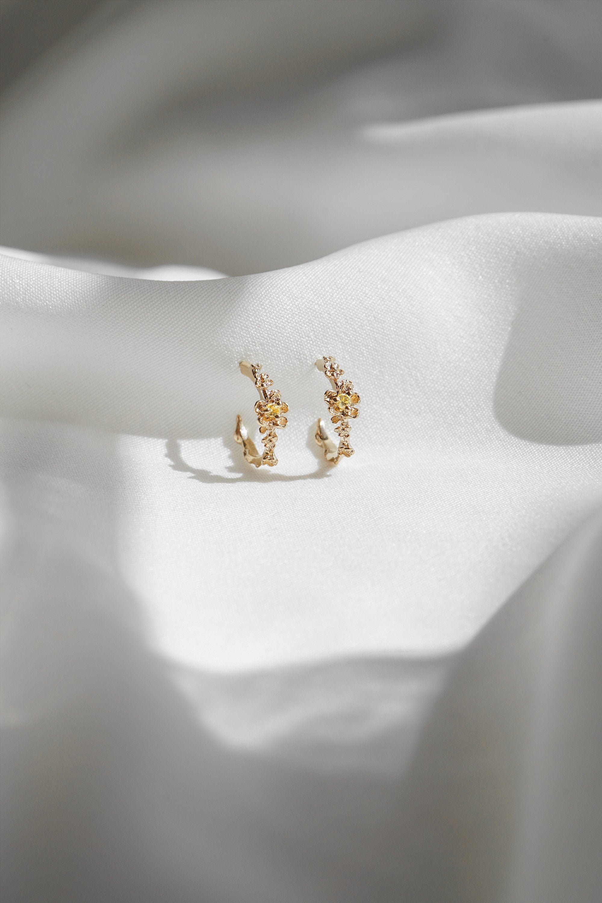 A pair of Laurie Fleming Jewellery "Small Buttercup Hoop" earrings, featuring petite hand-carved buttercup flowers with a yellow sapphire centre. The earrings are displayed against rippling smooth white fabric.
