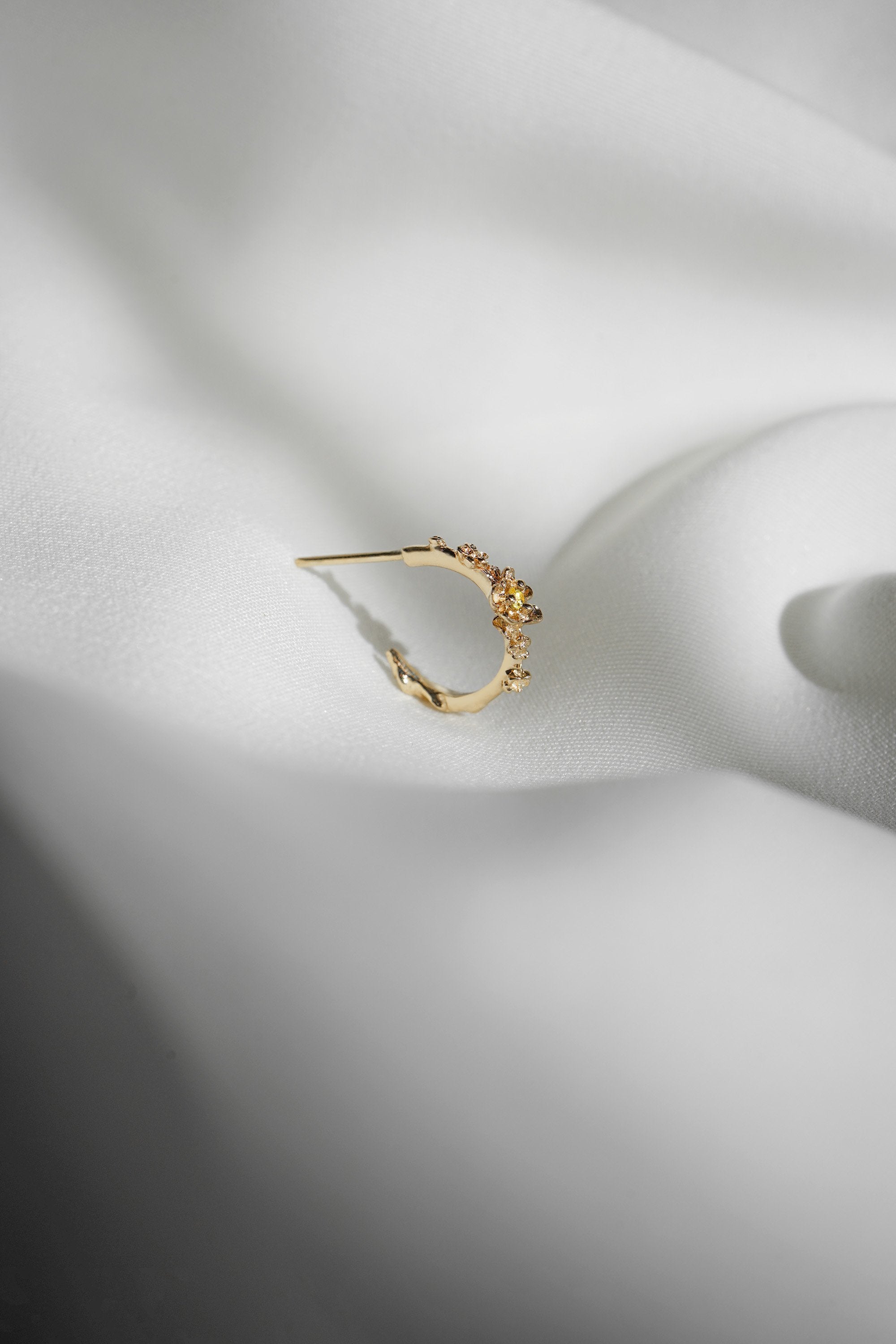 A single Laurie Fleming Jewellery "Small Buttercup Hoop" earring, seen from the side, featuring petite hand-carved buttercup flowers with a yellow sapphire centre. The earring is displayed against rippling smooth white fabric.