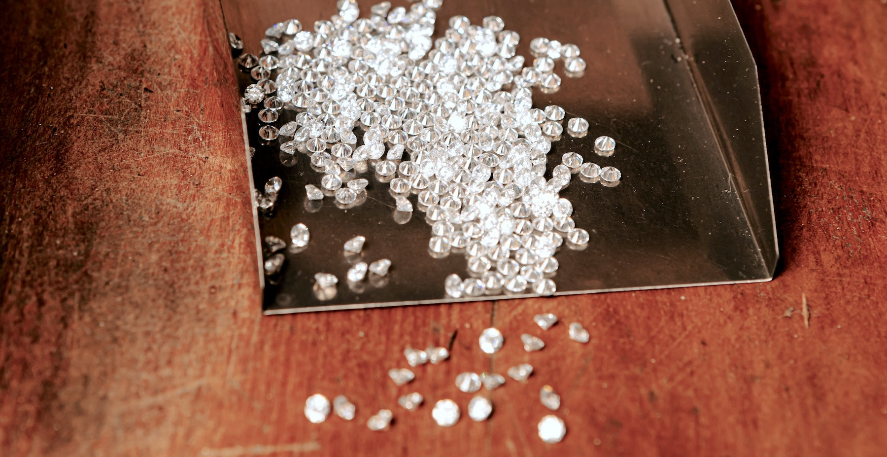 A stock image of a wooden table top. Upon it, there is a metal tray that contains a pile of tiny faceted diamonds, which spill out onto the table.
