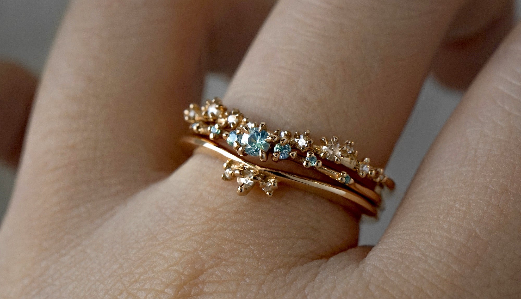 A hand wearing three rings stacked together. From top to bottom: White Wisp Band, Sapphire Water Lily Ring, Dewdrop Band.