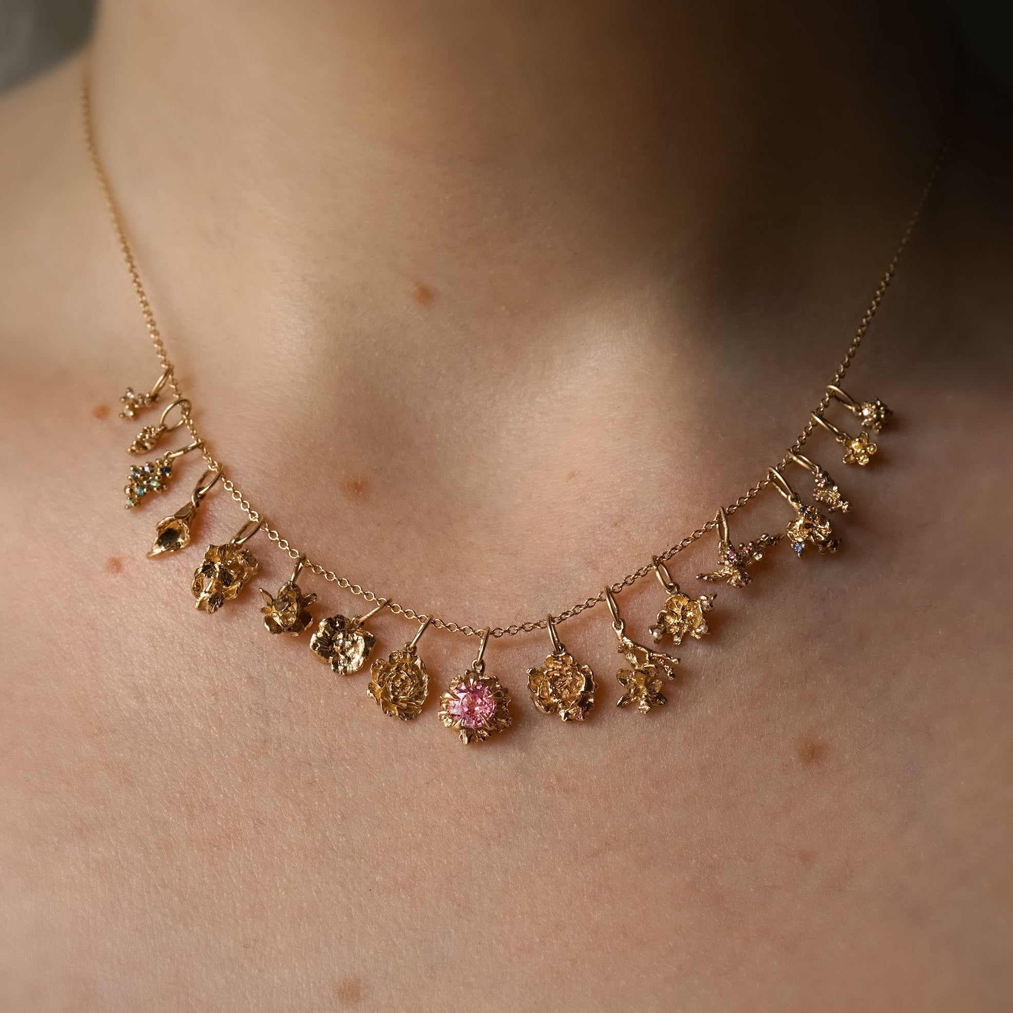 A close-up of a neck wearing a fine, 16 inch cable chain. On the chain are 17 handmade flower charms in solid gold with natural diamonds and sapphires, by Laurie Fleming Jewellery.