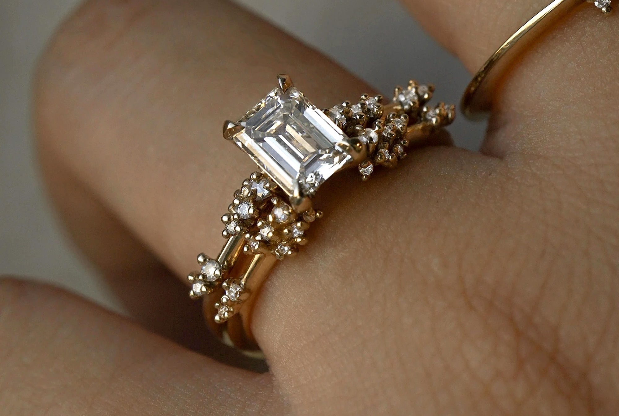a close-up of a hand wearing two rings together. On top, a "Emerald Cut Diamond Daphne Ring, Supreme", featuring a 1ct emerald cut diamond, and below, a "Daphne Band" open cuff ring. Both rings have rose cut and round brilliant cut diamonds on the band.
