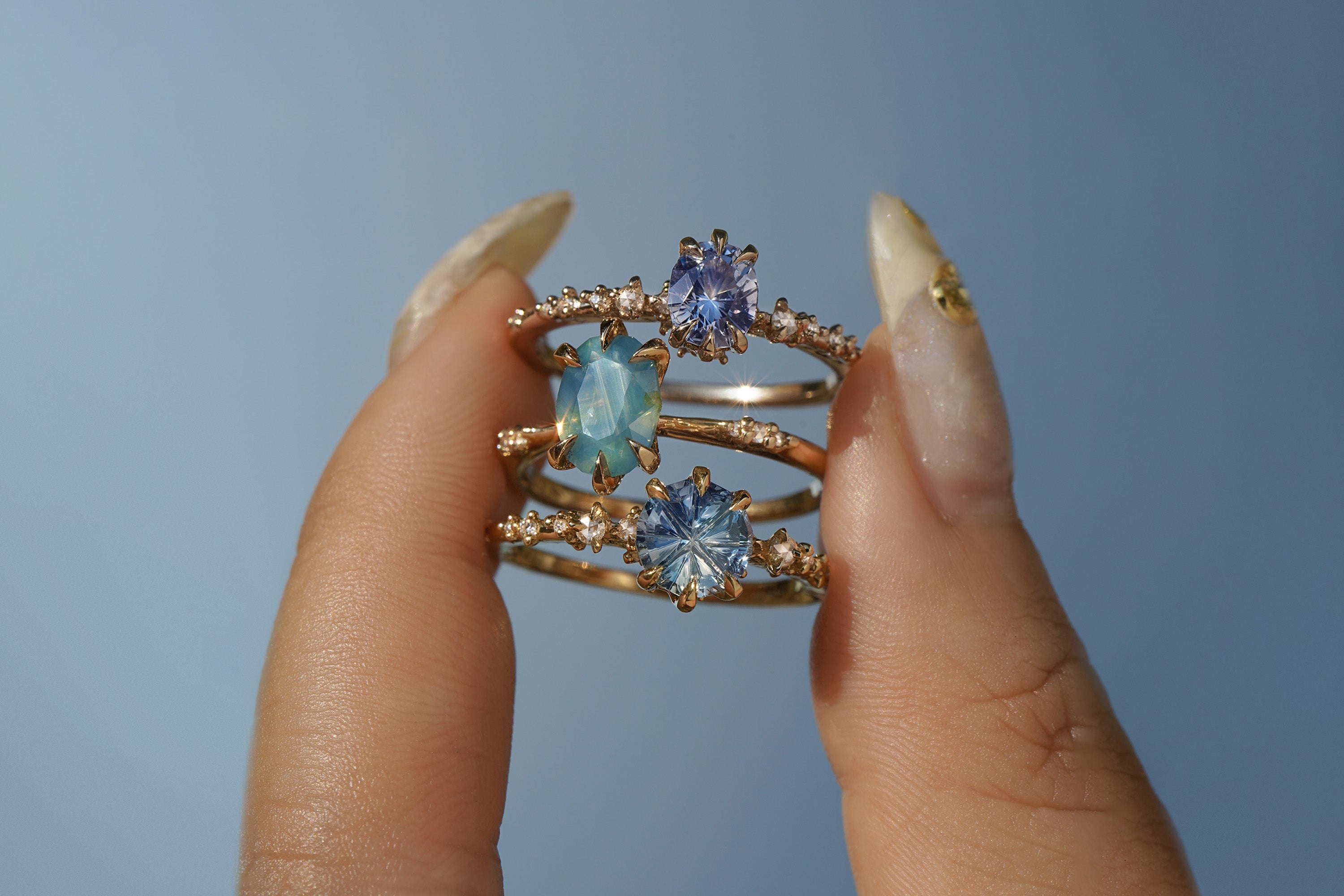 A group of three one of a kind rings by Laurie Fleming Jewellery. The rings are held between index finger and thumb and are held up against a bright blue sky.