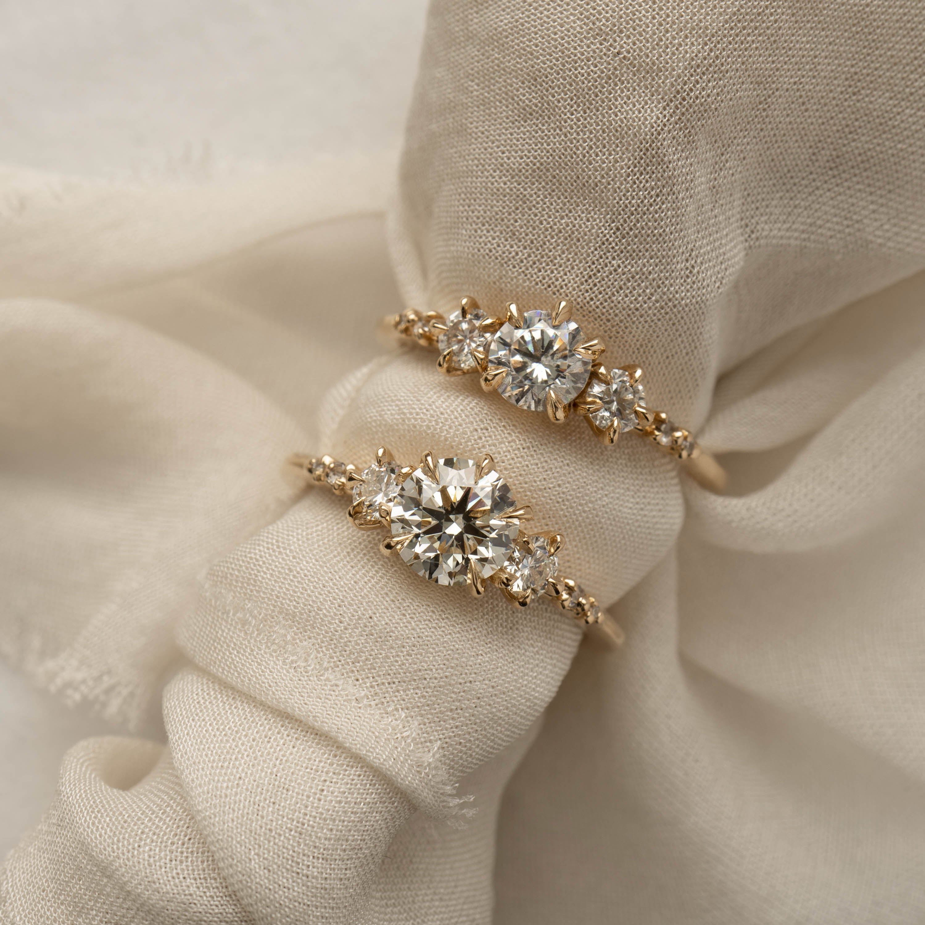 Comparison of two Brilliant Diamond Wild Iris Rings by Laurie Fleming Jewellery. Both rings are placed on a roll of fabric. The top ring features a half carat diamond centre, and the bottom ring features a one carat diamond centre. 