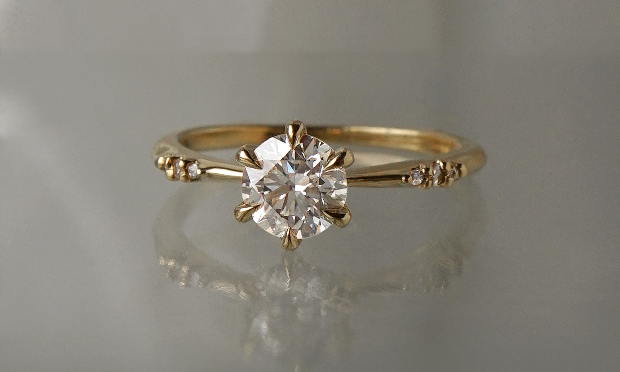 A delicate hand-carved one of a kind "Ilona" style engagement ring by Laurie Fleming Jewellery. The Ring features a 0.8ct round brilliant cut diamond, a tapered knife-edge band, and small diamond accents on the band shoulders.  The ring is situated against a medium grey background.