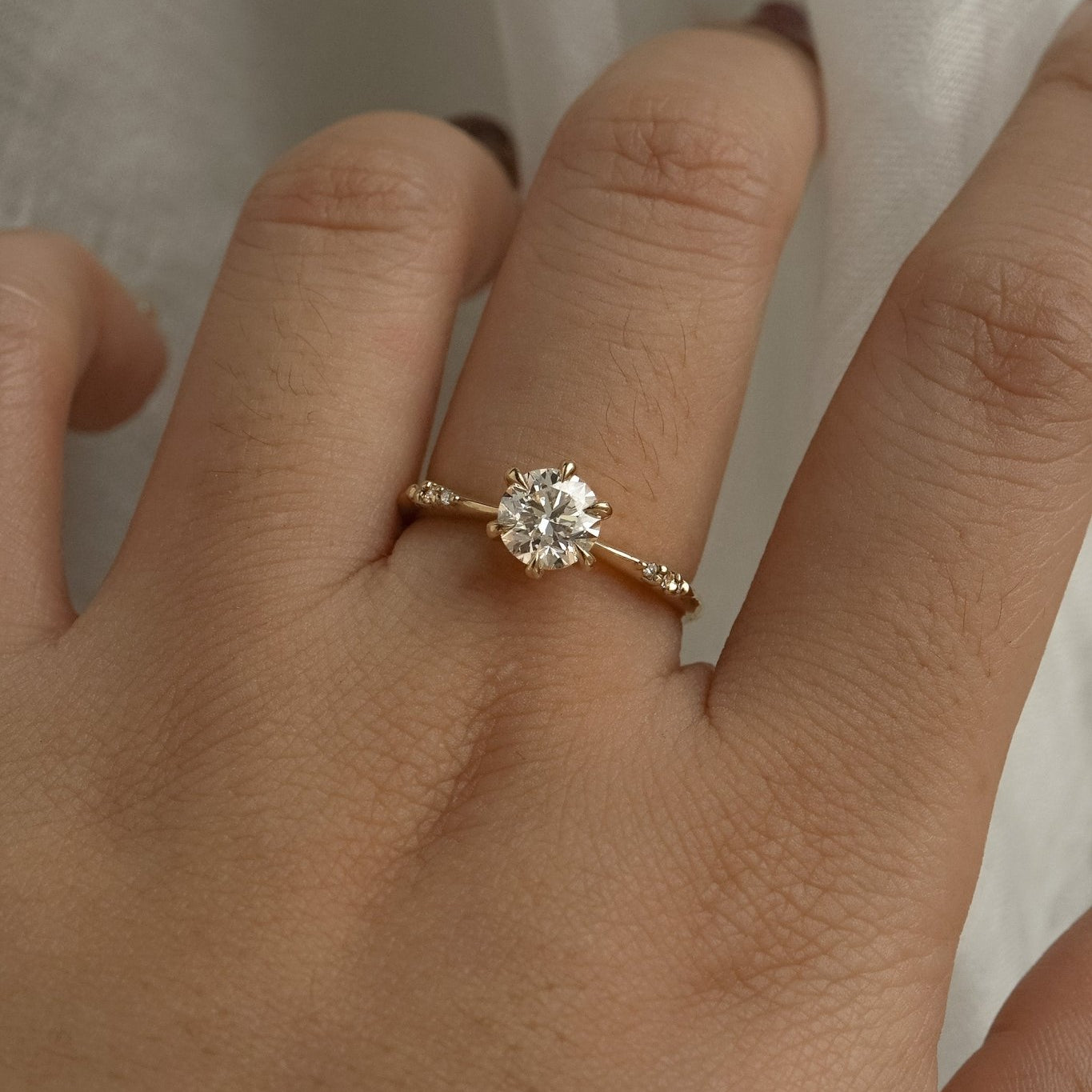 A delicate hand-carved one of a kind "Ilona" style engagement ring by Laurie Fleming Jewellery. The Ring features a 0.8ct round brilliant cut diamond, a tapered knife-edge band, and small diamond accents on the band shoulders.  The ring is worn on a hand posed in front of some draped gossamer silk fabric.