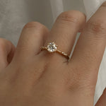 A delicate hand-carved one of a kind "Ilona" style engagement ring by Laurie Fleming Jewellery. The Ring features a 0.8ct round brilliant cut diamond, a tapered knife-edge band, and small diamond accents on the band shoulders.  The ring is worn on a hand posed in front of some draped gossamer silk fabric.