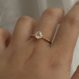 A delicate hand-carved one of a kind "Ilona" style engagement ring by Laurie Fleming Jewellery. The Ring features a 0.8ct round brilliant cut diamond, a tapered knife-edge band, and small diamond accents on the band shoulders.  The ring is worn on a hand posed in front of some draped gossamer silk fabric.