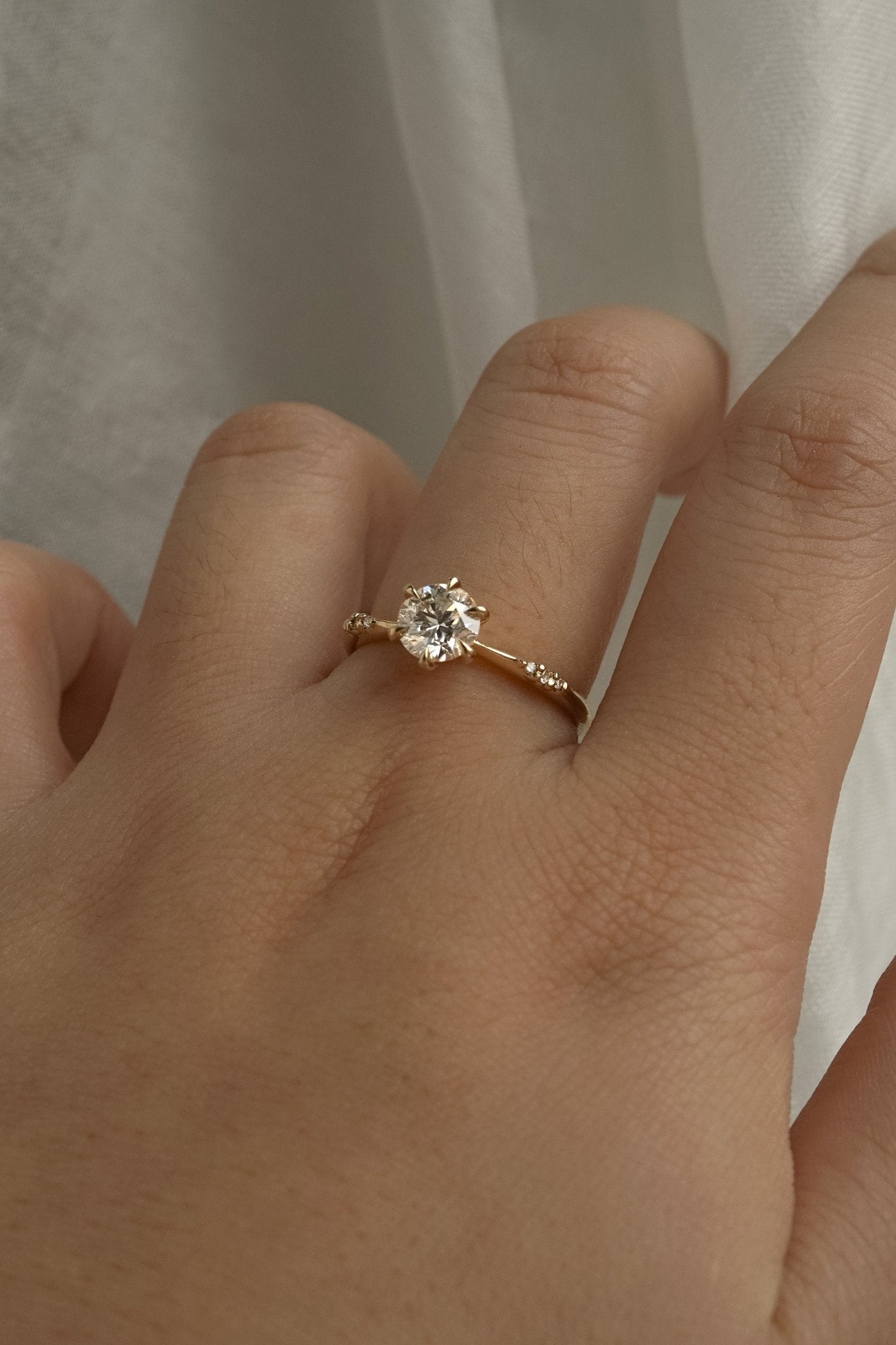 A delicate hand-carved one of a kind "Ilona" style engagement ring by Laurie Fleming Jewellery. The Ring features a 0.8ct round brilliant cut diamond, a tapered knife-edge band, and small diamond accents on the band shoulders.  The ring is worn on a hand posed in front of some draped gossamer silk fabric.