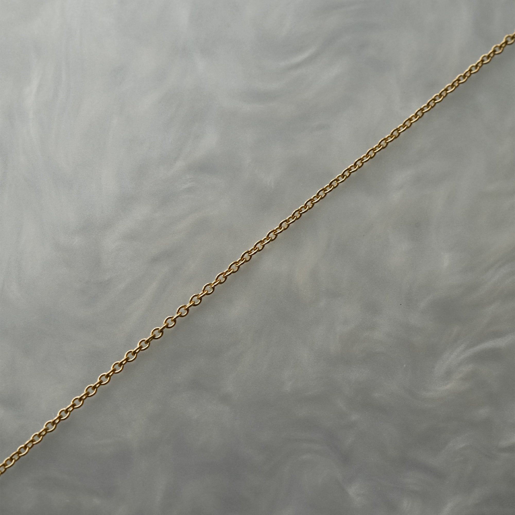 1mm fine and delicate cable chain with smooth oval links in solid 14k gold, perfect for wearing on its own or with charms or pendants. The chain is against a grey background with swirling patterns.