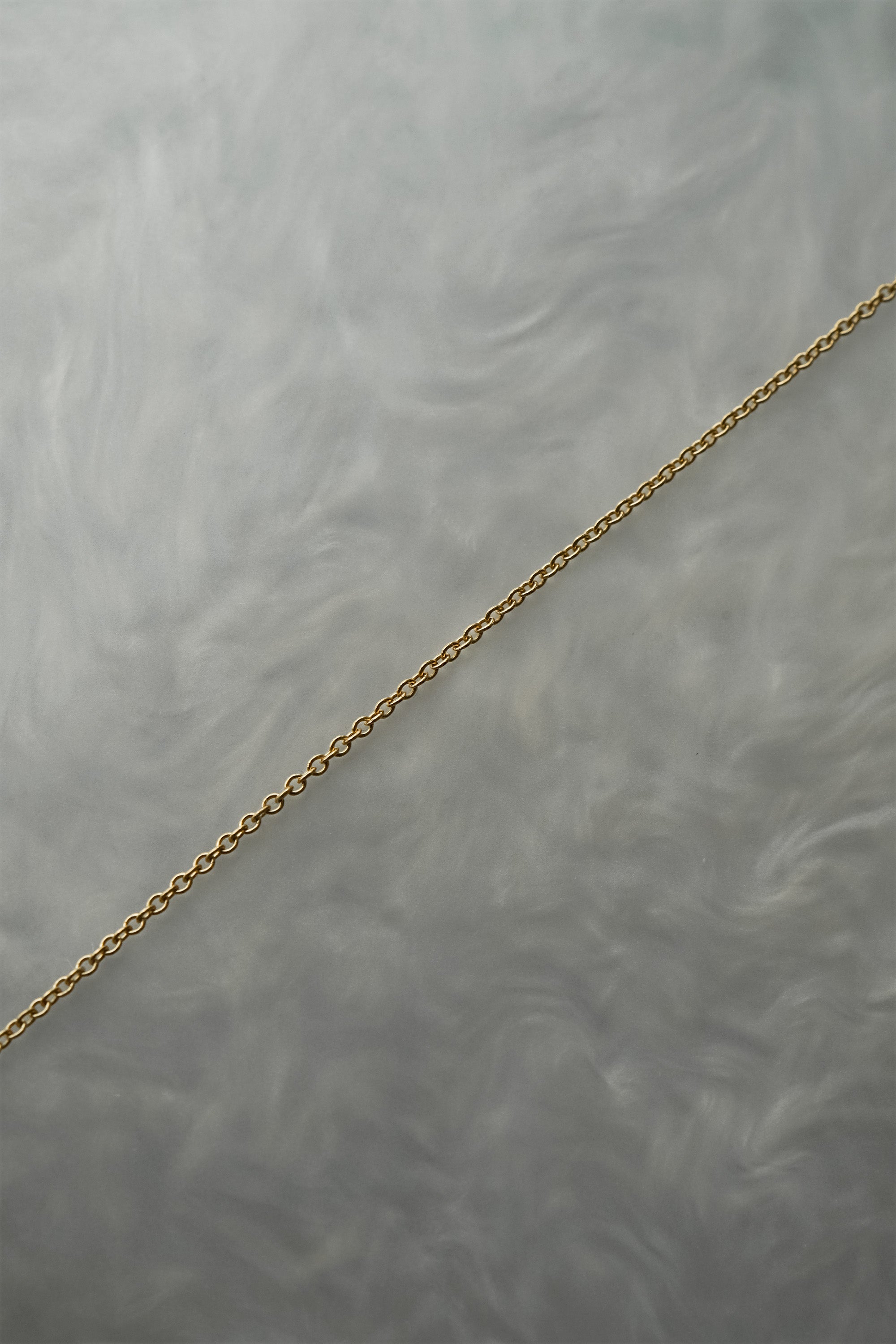 1mm fine and delicate cable chain with smooth oval links in solid 14k gold, perfect for wearing on its own or with charms or pendants. The chain is against a grey background with swirling patterns.