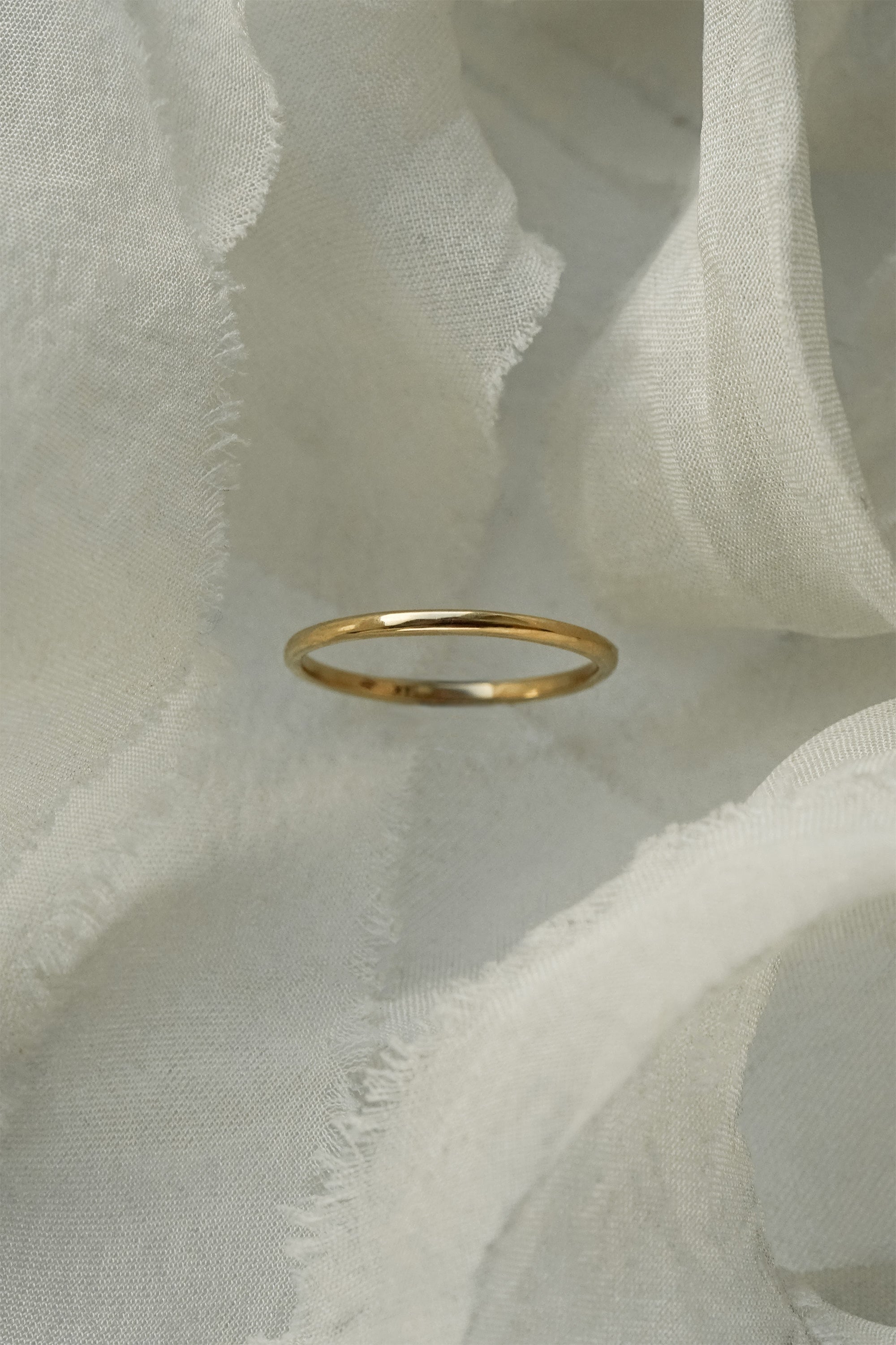 A stock photo of Laurie Fleming Jewellery's plain solid gold "Myrtle" band, a 1.4mm wide stacking ring/wedding band, handmade in Toronto. The ring is standing on its own surrounded by pieces of raw white silk.