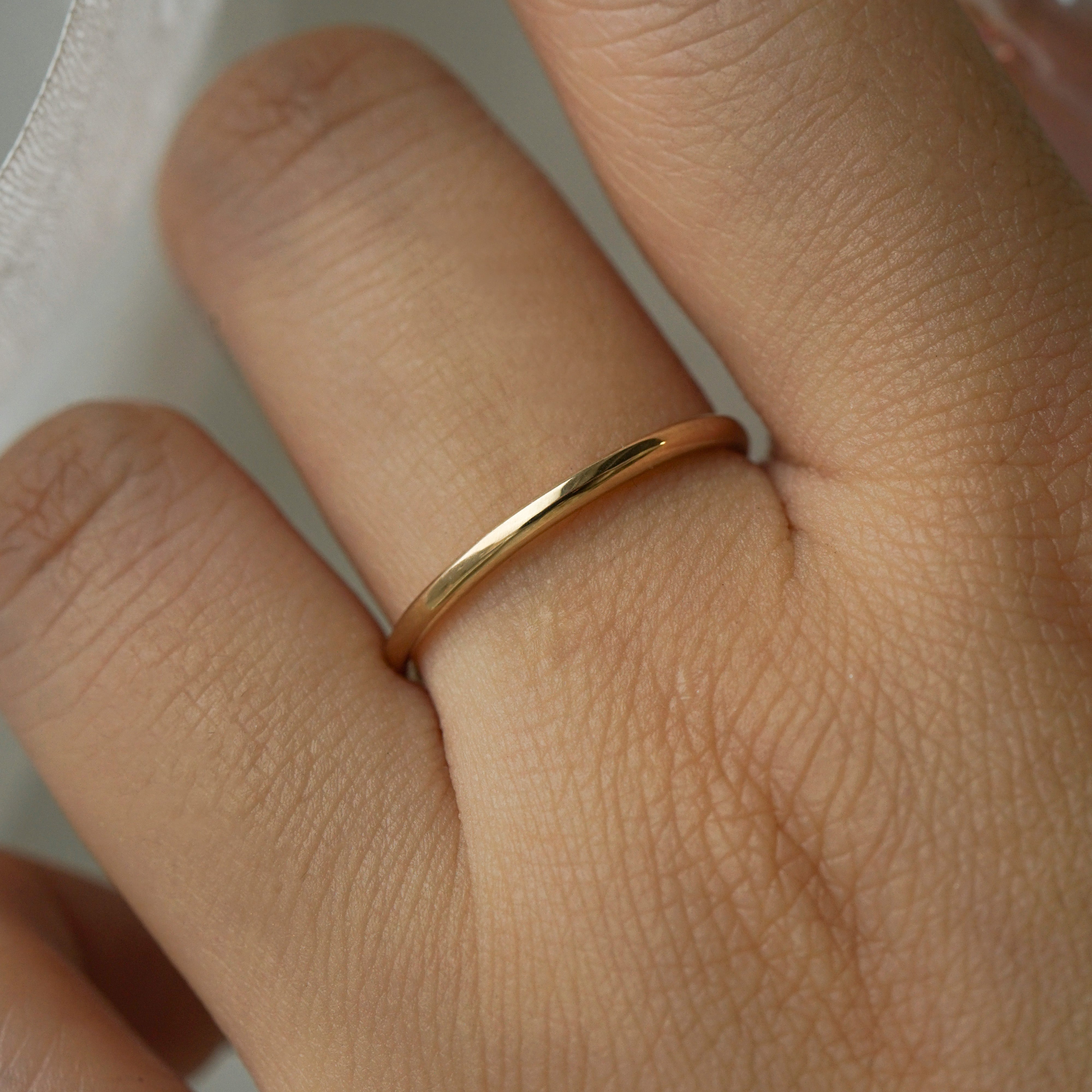 A stock photo of Laurie Fleming Jewellery's plain solid gold "Myrtle" band, a 1.4mm wide stacking ring/wedding band, handmade in Toronto. The ring is worn on a hand with a light grey background.