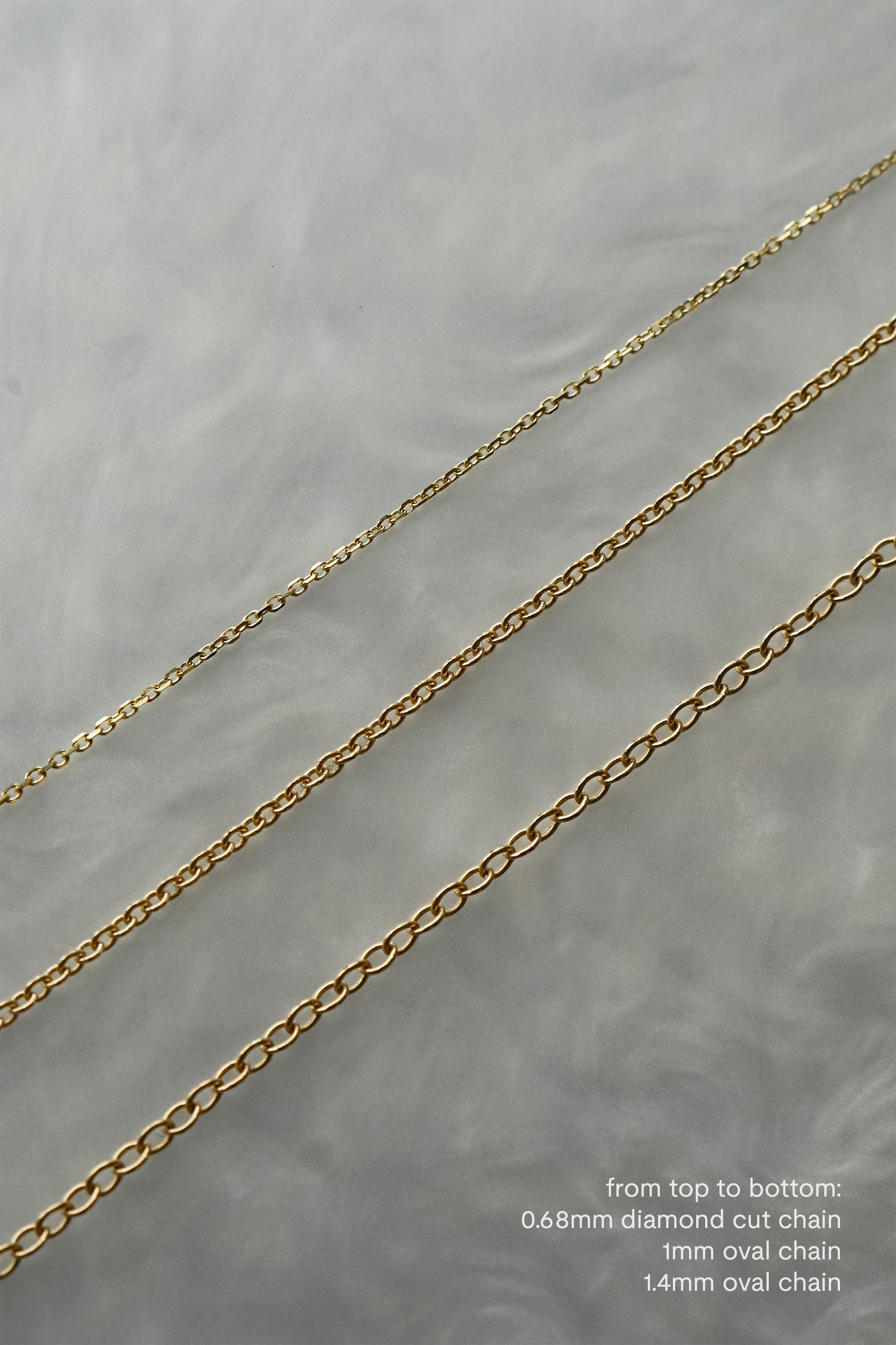 Three delicate solid gold chains laid out on a grey background with swirling patterns. From top to bottom: 0.68mm diamond-cut chain, 1mm petite smooth oval chain, 1.4mm smooth oval chain.