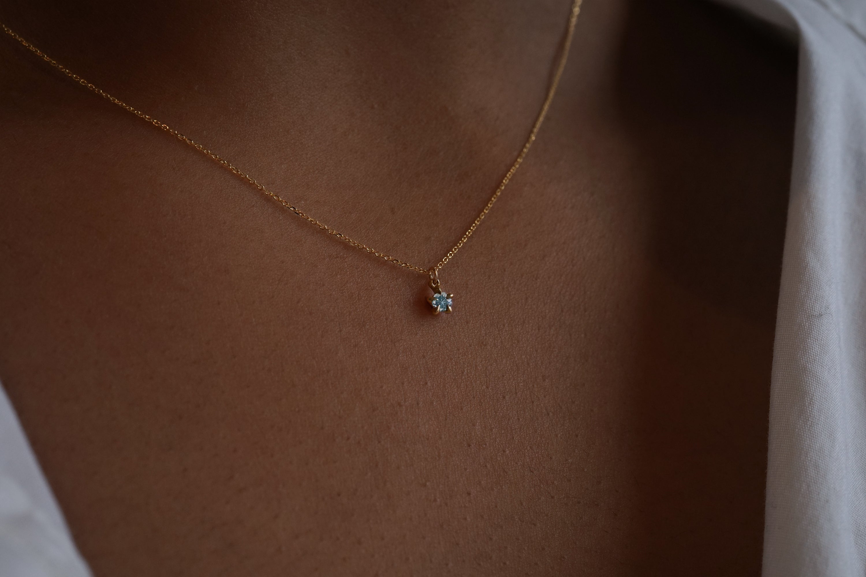 A close up shot of the blue-green teal sapphire Moondew pendant necklace by Laurie Fleming Jewellery in yellow gold, modelled on dark brown skin.